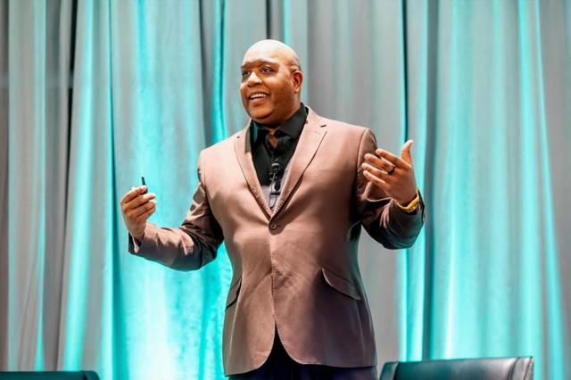 Omar L. Harris delivering a keynote on stage at a leadership event, speaking to a seated audience with a presentation screen behind him